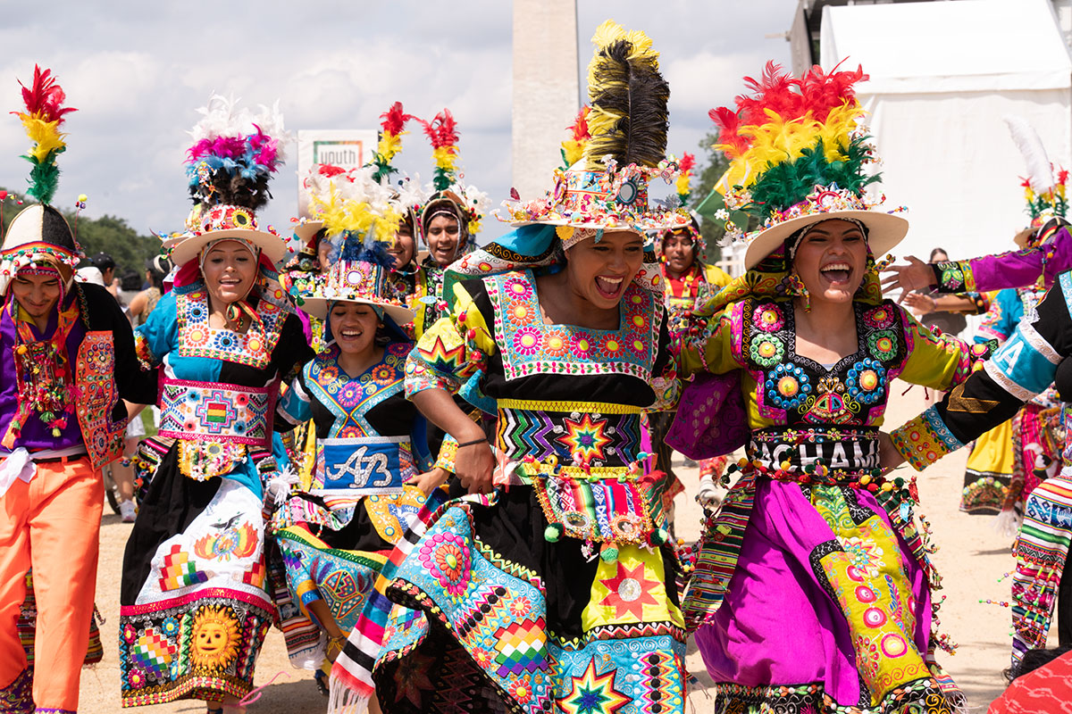 A group of smiling performers wearing colorful, ornate traditional costumes with large feather headdresses dancing in a festival parade.