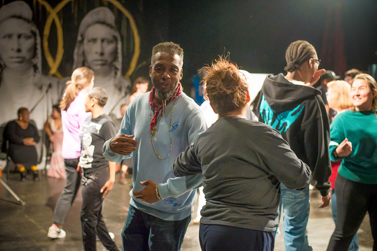Pairs of people do-si-do, with their arms linked at the elbows, in an indoor dance space.