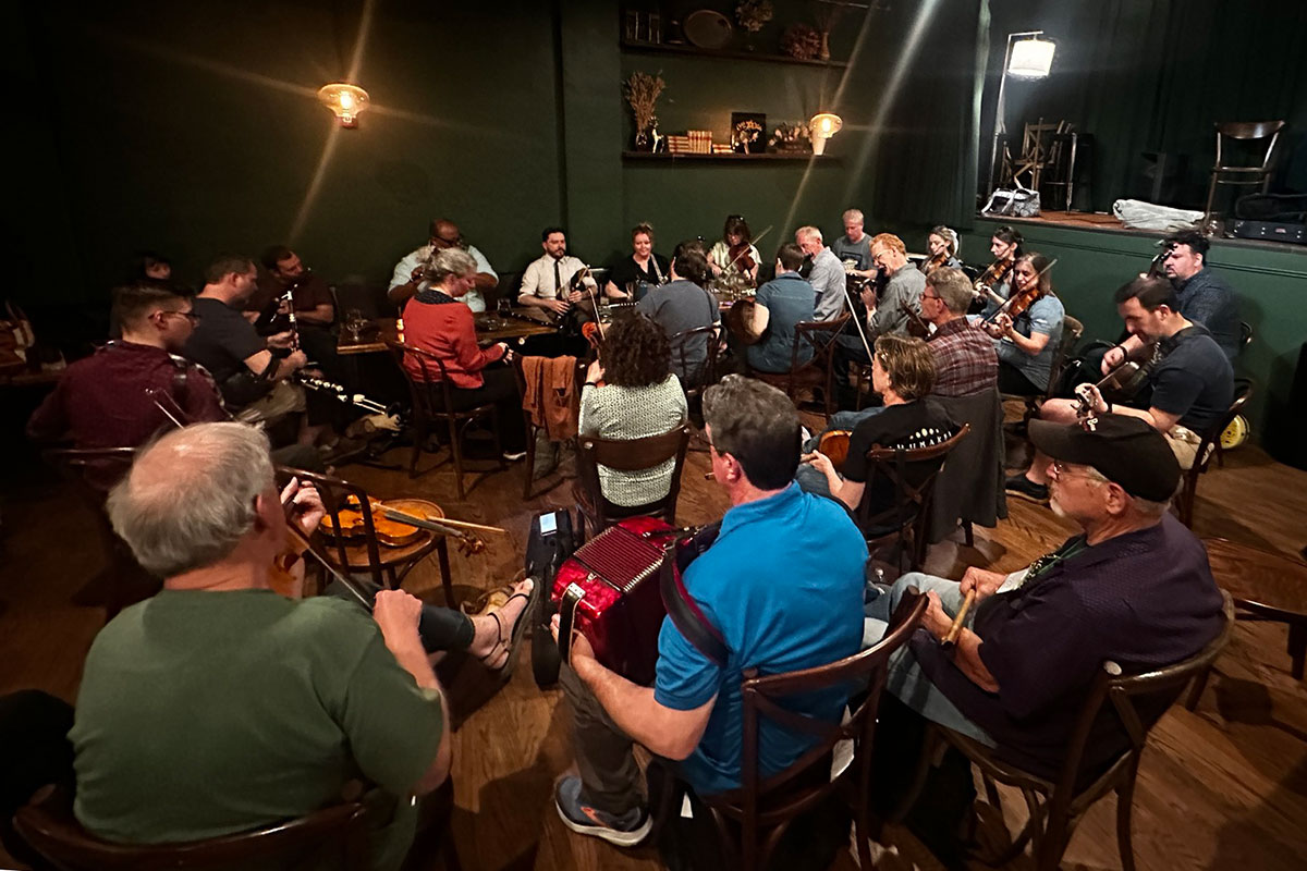 Two dozen people play various musical instruments, seated in loose concentric circles in a large room. There are fiddles, an accordion, guitars, and clarinets.