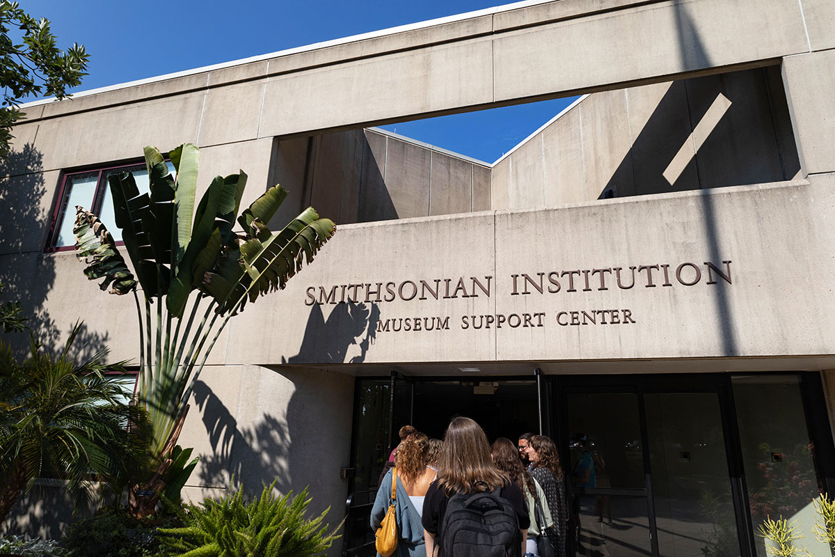 A group of people walk into the open doors of a concrete building, with words above the doorway: Smithsonian Institution Museum Support Center.