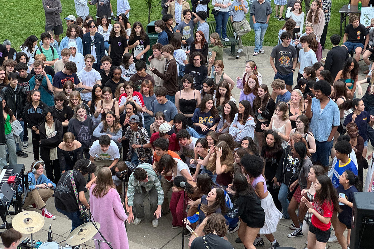 From above, a crowd of a teenage students gathers in front of a band playing on an outdoor stage.