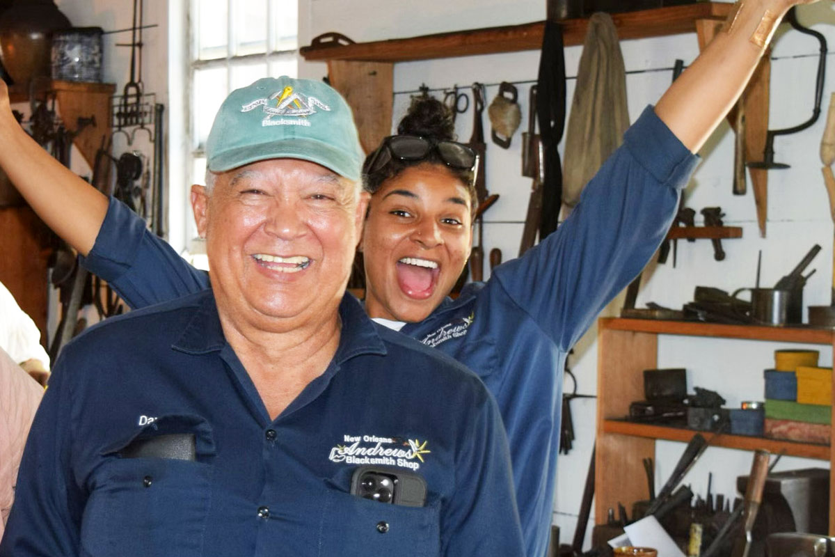An elder man and a young woman in matching navy blue work shirts smile for the camera. The woman, a step behind the man, smiles widely with both arms raised high. Tools hang on the wall behind them.