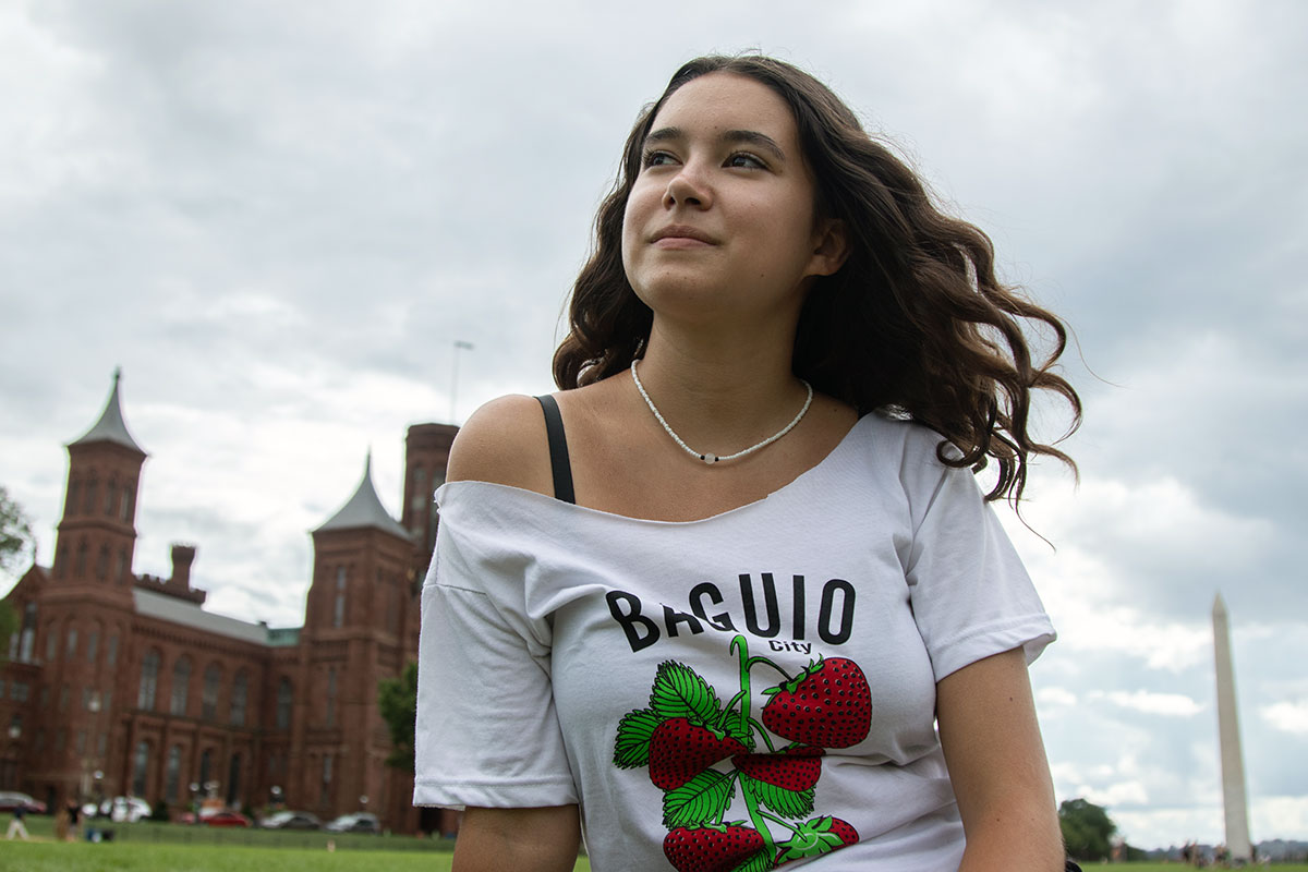A teen girl with long wavy brown hair and a slight smile poses sitting in the grass on the National Mall, with the red-brick Smithsonian Castle and Washington Monument in the background.