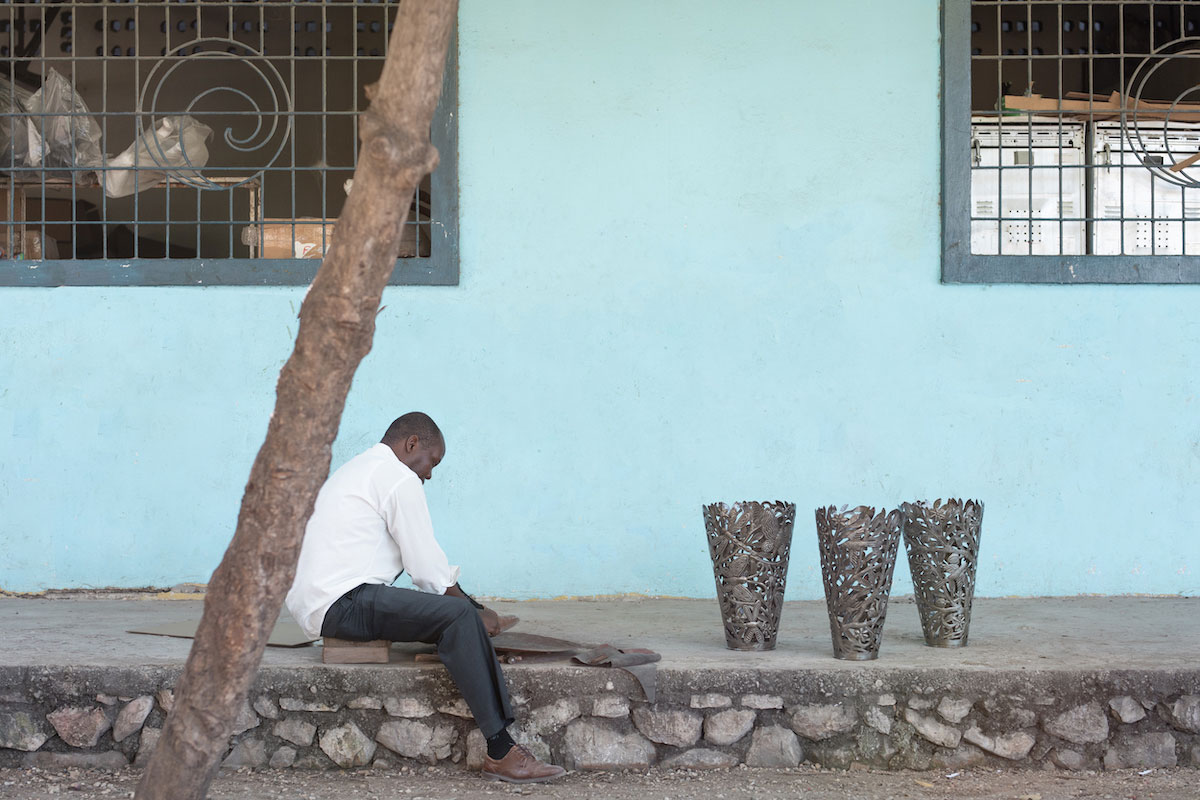 A Black man sits on a paved walkway, working a sheet of metal. Three decorative metal vases sit nearby.