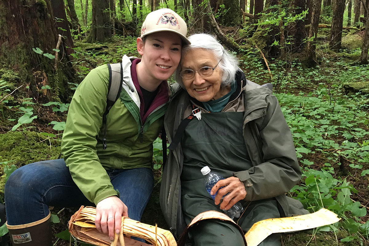 A young woman and an elder woman pose with their arms around each other, seated in a forest. They both have in their laps strips of cedar bark.