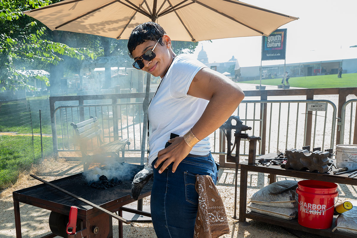 A young woman with short dark hair, dark sunglasses, and a gold cuff bracelet, with a handkerchief sticking out of her back pocket, tends to a smoking iron forge. She turns back to smile at the camera. Behind her, the Capitol Building and a sign for Youth and the Future of Culture on the National Mall.