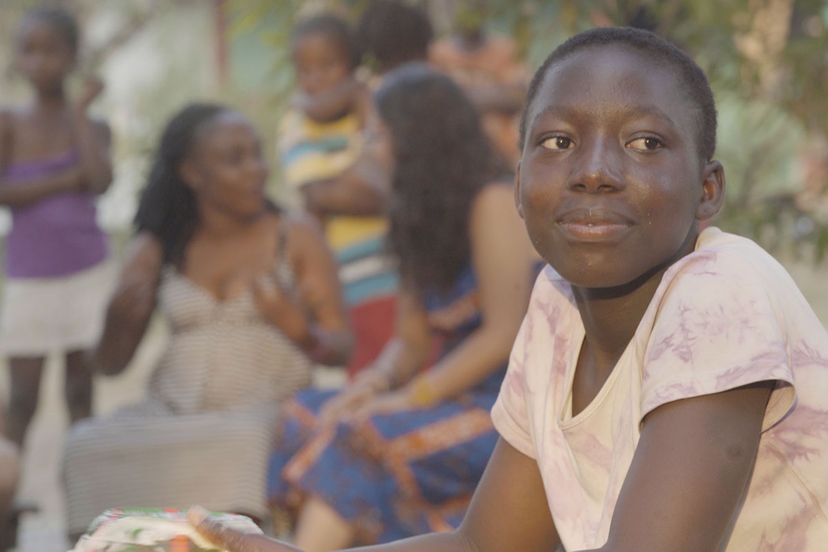 Film still of a person sitting outside, looking into the distance, while others gather behind them chatting.