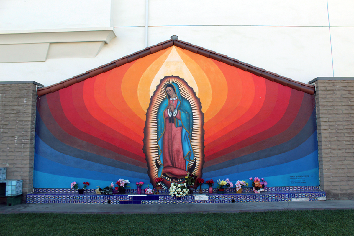 A mural on an outdoor wall, with a virgin figure at the center, with orange, red, purple, and blue stripes radiating out from her. At the base of the mural, several bouquets of flowers on a tiled step.