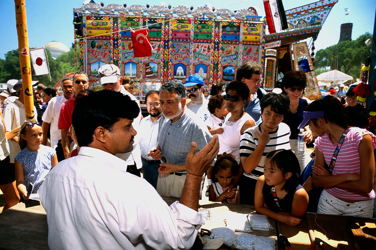A man gestures in front of a crowd of kids and adults. In the background, a box truck that has been elaborately painted and adorned in bright colors.