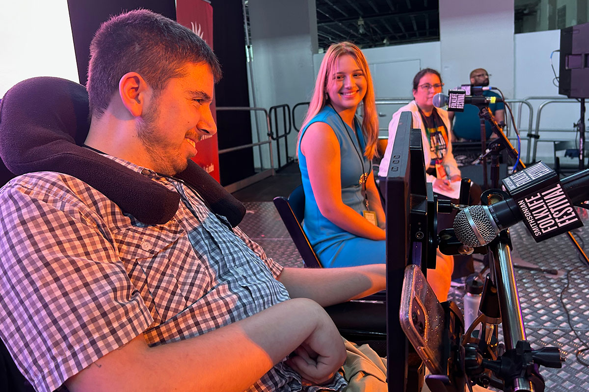 Three people sit on an illuminated stage. In the foreground, a young man in a plaid shirt smiles toward the audience. In front of him is a communication devide with a screen and a Folklife Festival microphone. The young woman next to him turns to look at 