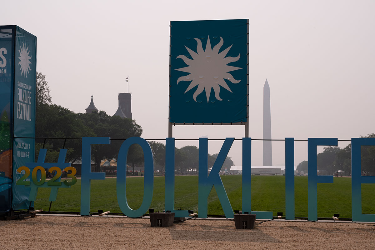 In the foreground, large block cutout letters reading #2023FOLKLIFE, under a banner with the Smithsonian sun logo. In the background, the Smithsonian Castle and Washington Monument.