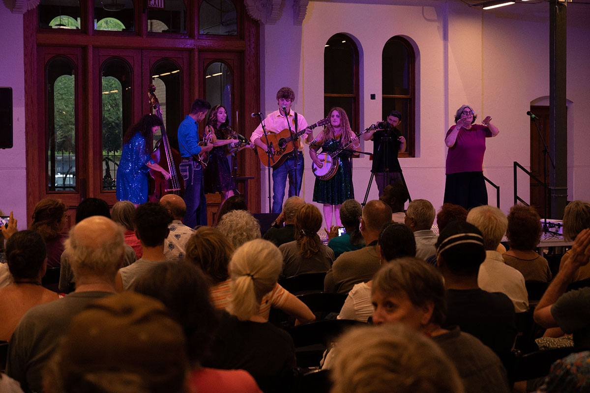 On stage, a five-piece string band perform in front of a crowd. Also on stage are a videographer and an ASL interpreter.