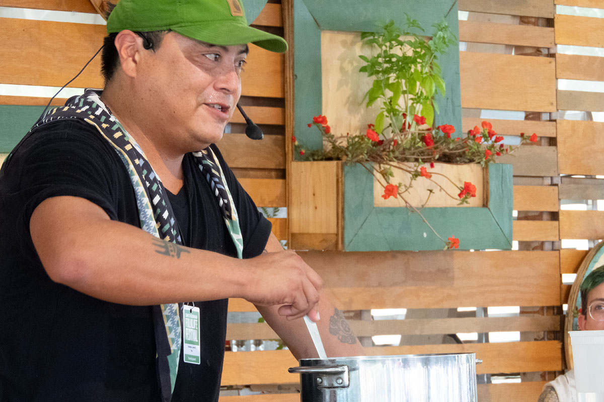 A man in green baseball cap and black shirt, wearing a headset microphone, stirs a put in an outdoor kitchen.