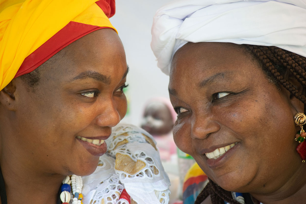 Two women share a glance and a smile with each other. They both wear headwraps.