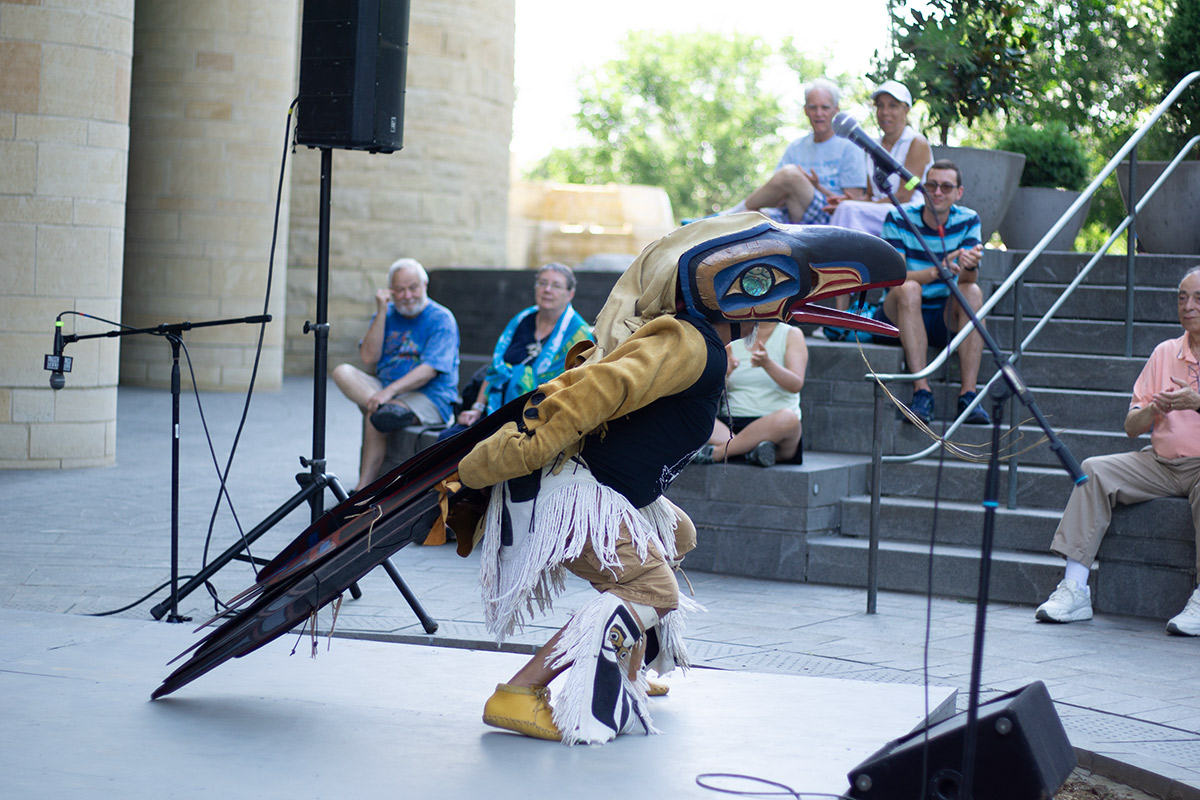 A man wearing birdlike regalia stands in front of an audience outdoors.