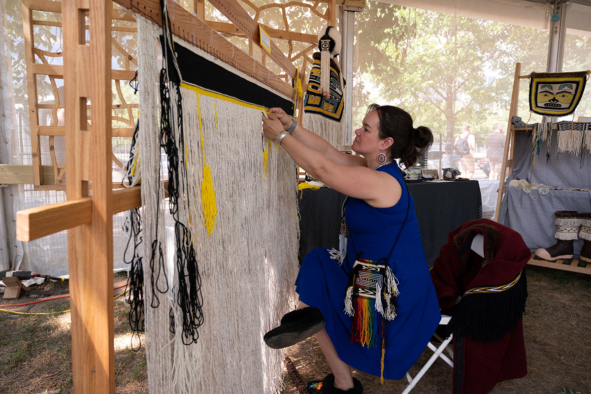 A woman sits in a wooden loom under a tent outdoors.
