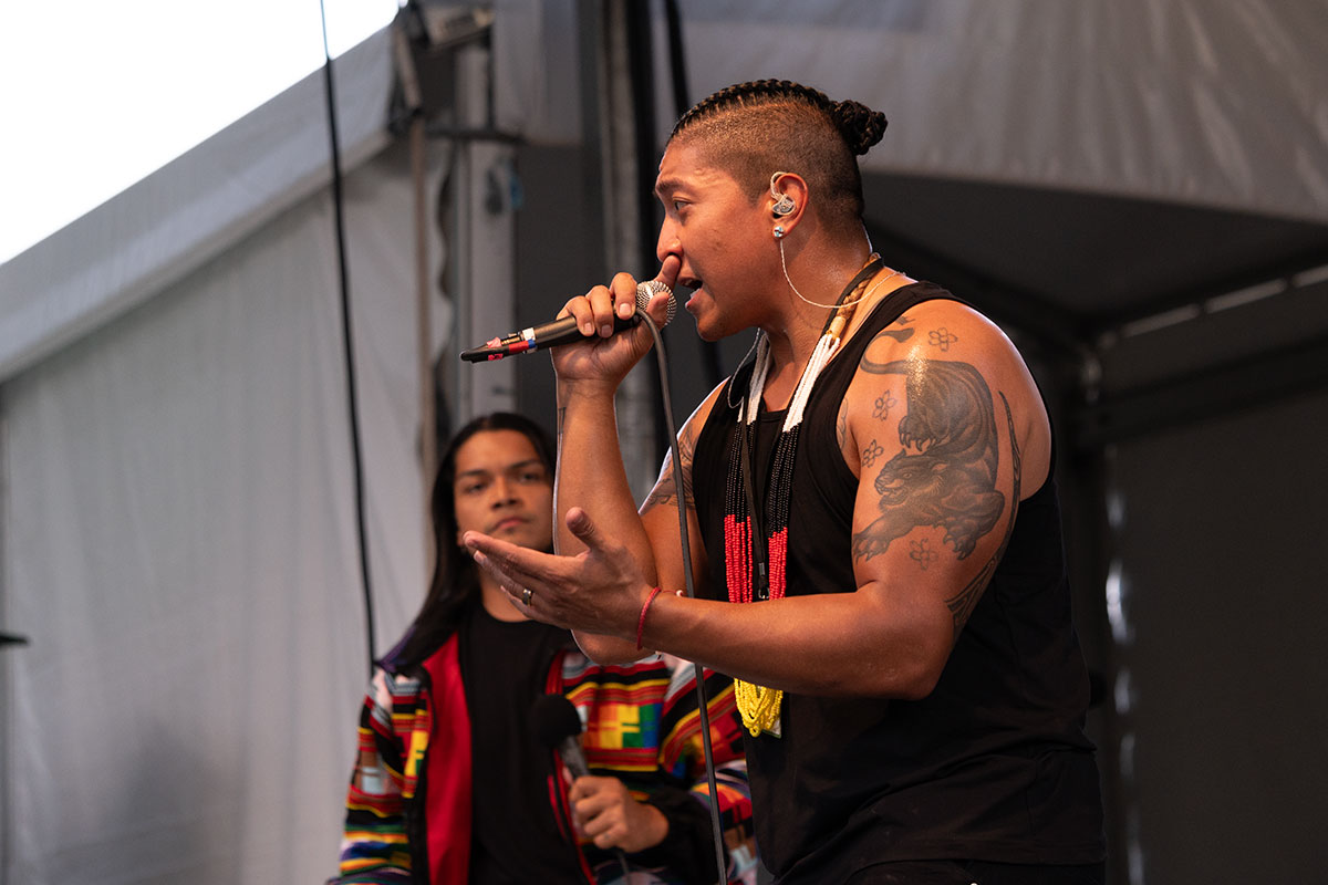 Two people stand on stage, one facing the other, who faces the audience. Both have microphones in hand, and the one facing the audience raps into it. 