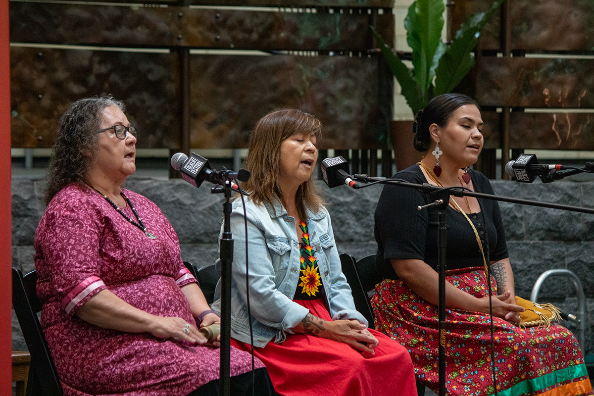 Three women sitting on a stage, singing into microphones on stands.
