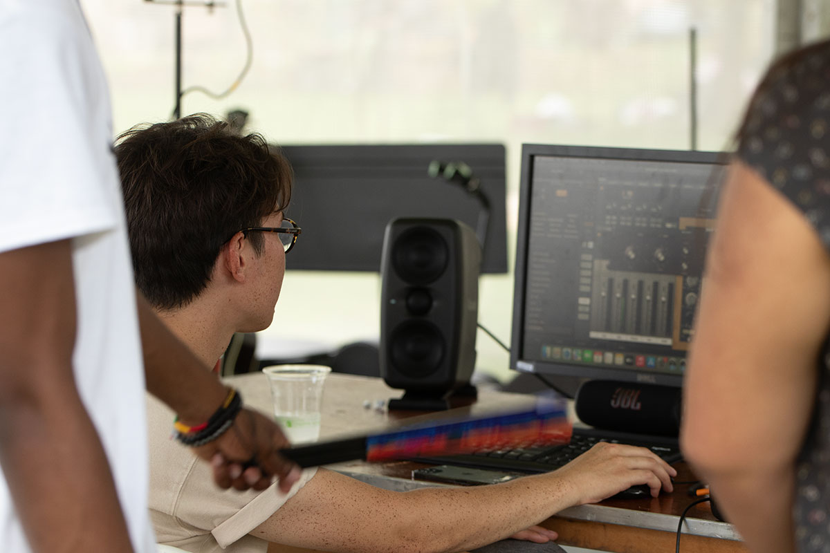 A man works on a digital audio workstation on a computer, set up under a tent outdoors. Two people, their heads out of frame, stand behind him to watch.