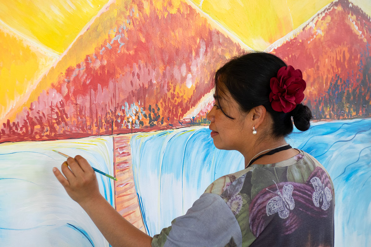 A woman with long dark hair in a low bun, adorned with a faux red rose, use a skinny paintbrush on a mural that takes up the full frame, depicting a pathway through a blue body of water, leading toward red mountains and a yellow sky.