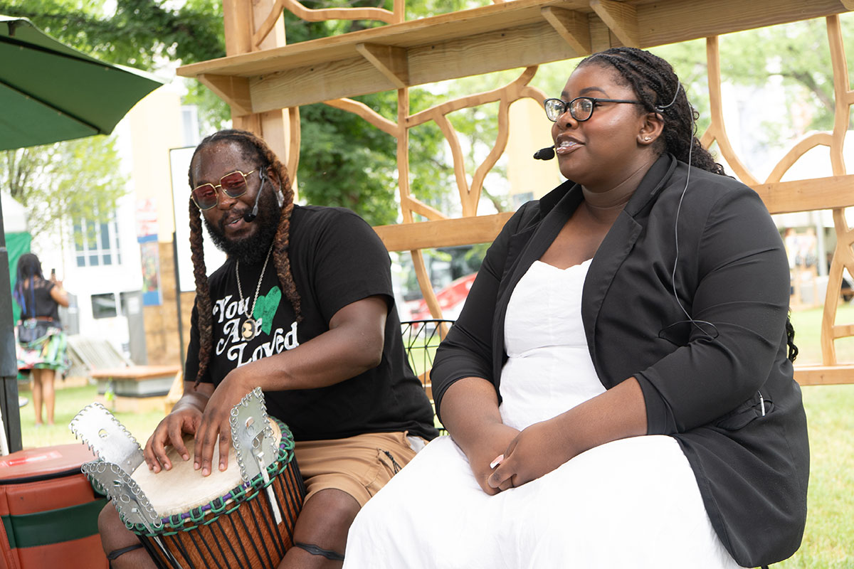 Two people sit and perform on an outdoor stage: a man with a dark beard, sunglasses, and two long braids on a hand drum with rectangular tin snares, and a woman in a white dress and black blazer with dark hair in braids. They both wear headset microphones