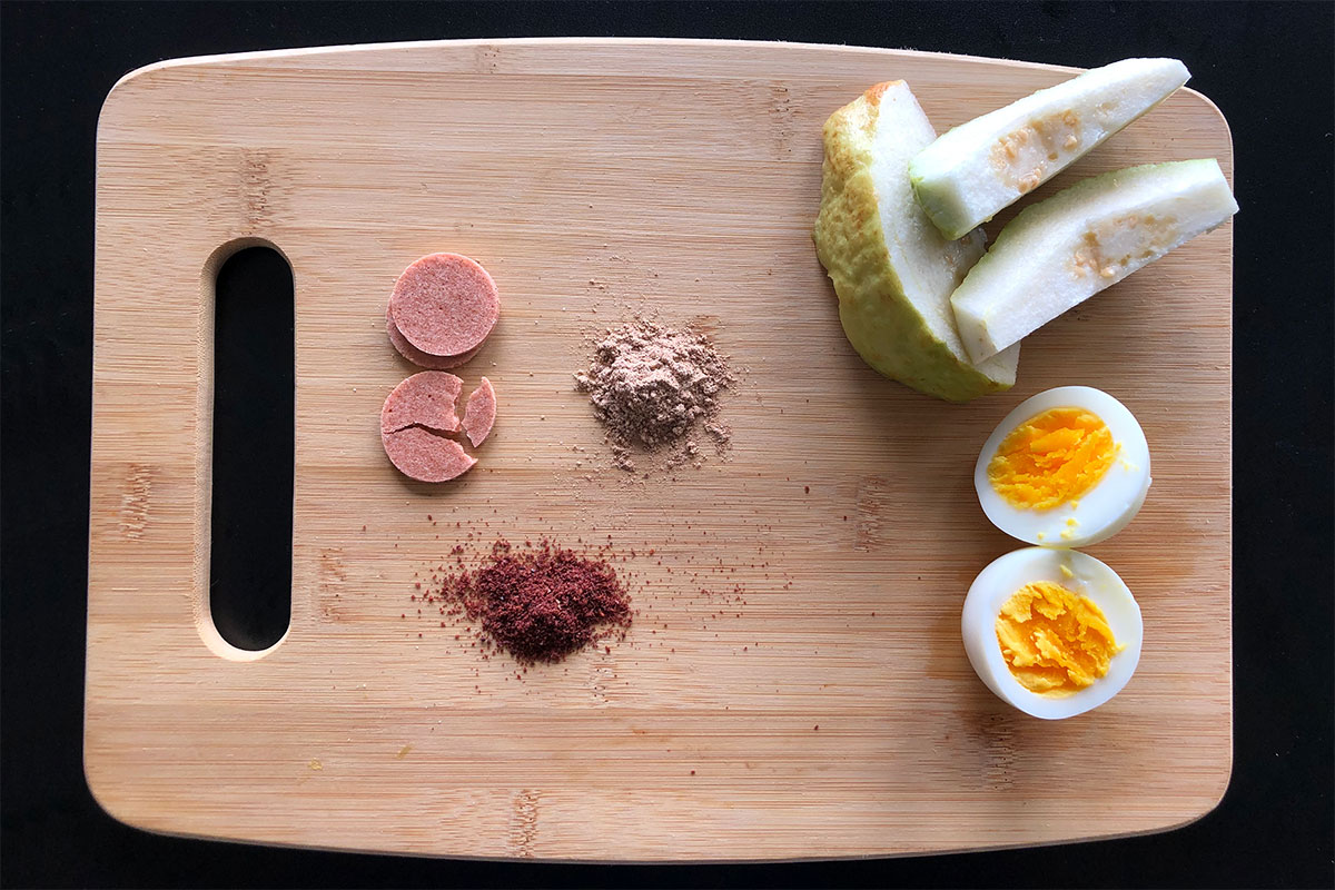 Cutting board containing a lineup of snacks: pink circles, green and white slices of guava, a hard-boiled egg, and two pinched of pinkish powders.