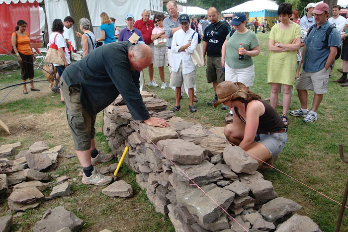 A man and a young woman work on either side of a low stone wall, with a crowd of people watching.