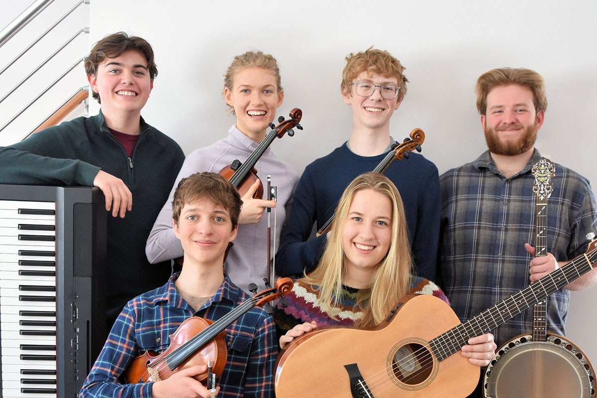 Six young people pose, smiling, in two rows with their instruments: electric keyboard, fiddles, banjo, and acoustic guitar.