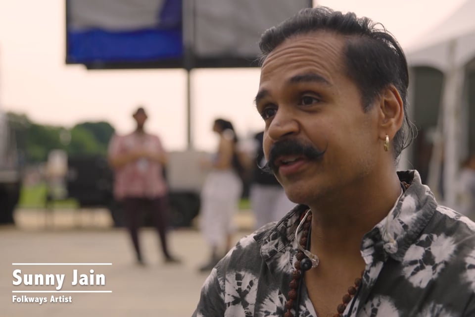 A South Asian man with black handlebar moustache, gold earring, and tie-dyed shirts speaks, seated. HIs name is in text: Sunny Jain, Folkways Artist. Behind him, backstage of an outdoor festival.