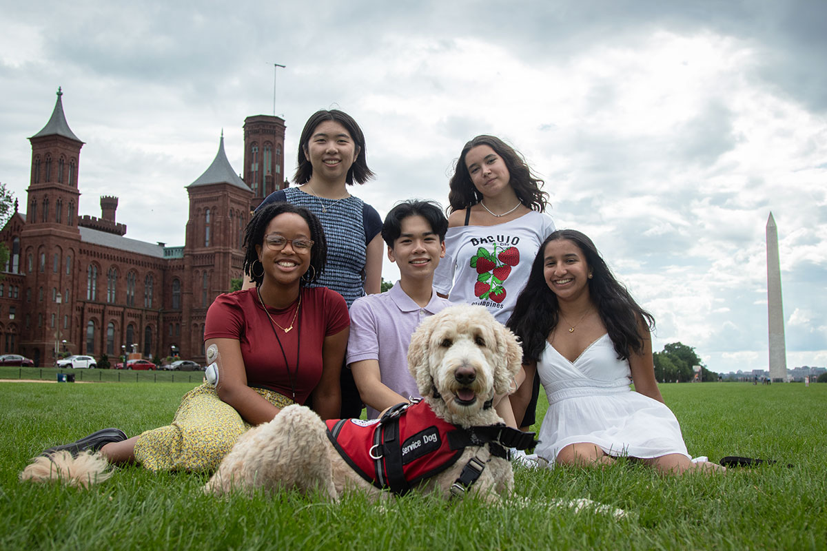 Five teens and one dog pose together, seated on the National Mall, with the Smithsonian Castle and Washington Monument behind them.