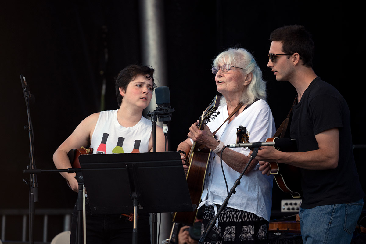 Three musicians gather around a microphone on an outdoor stage singing. They are each holding a string instrument.