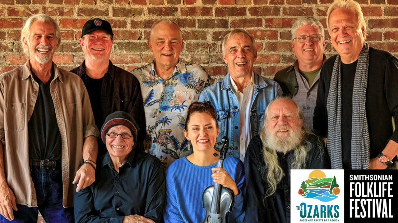 Nine musicians stand in front of a brick wall, a woman with a fiddle at the center.