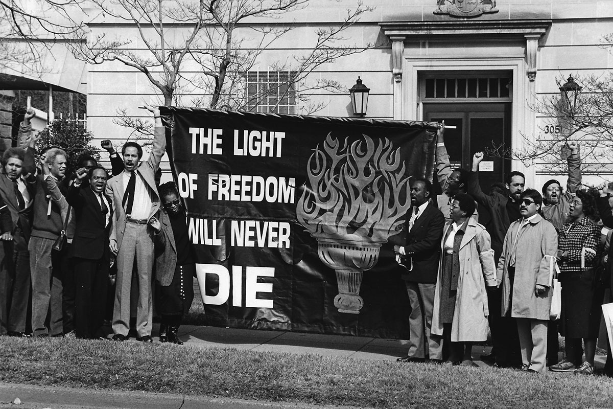 A group of mostly Black people gathered in protest in front of a government building, holding up a banner that says THE LIGHT OF FREEDOM WILL NEVER DIE. Black-and-white photograph.
