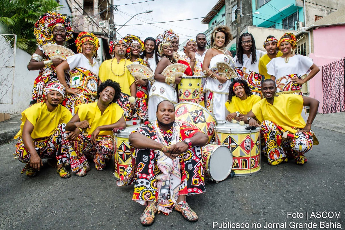 Photo of Ilê Aiyê, a group of eighteen people posing in the middle of street with decorated bass drums, bright yellow and white tops, brightly patterned matching pants, fans, and some with headdresses. In the background, palm fronds and pastel colored buildings.