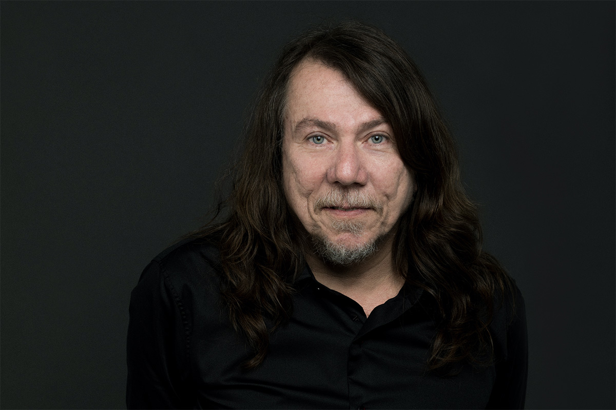 Photo of Lenine, with long brown hair, grayish goatee, and black dress shirt, smiling for the camera in front of a plain black backdrop.