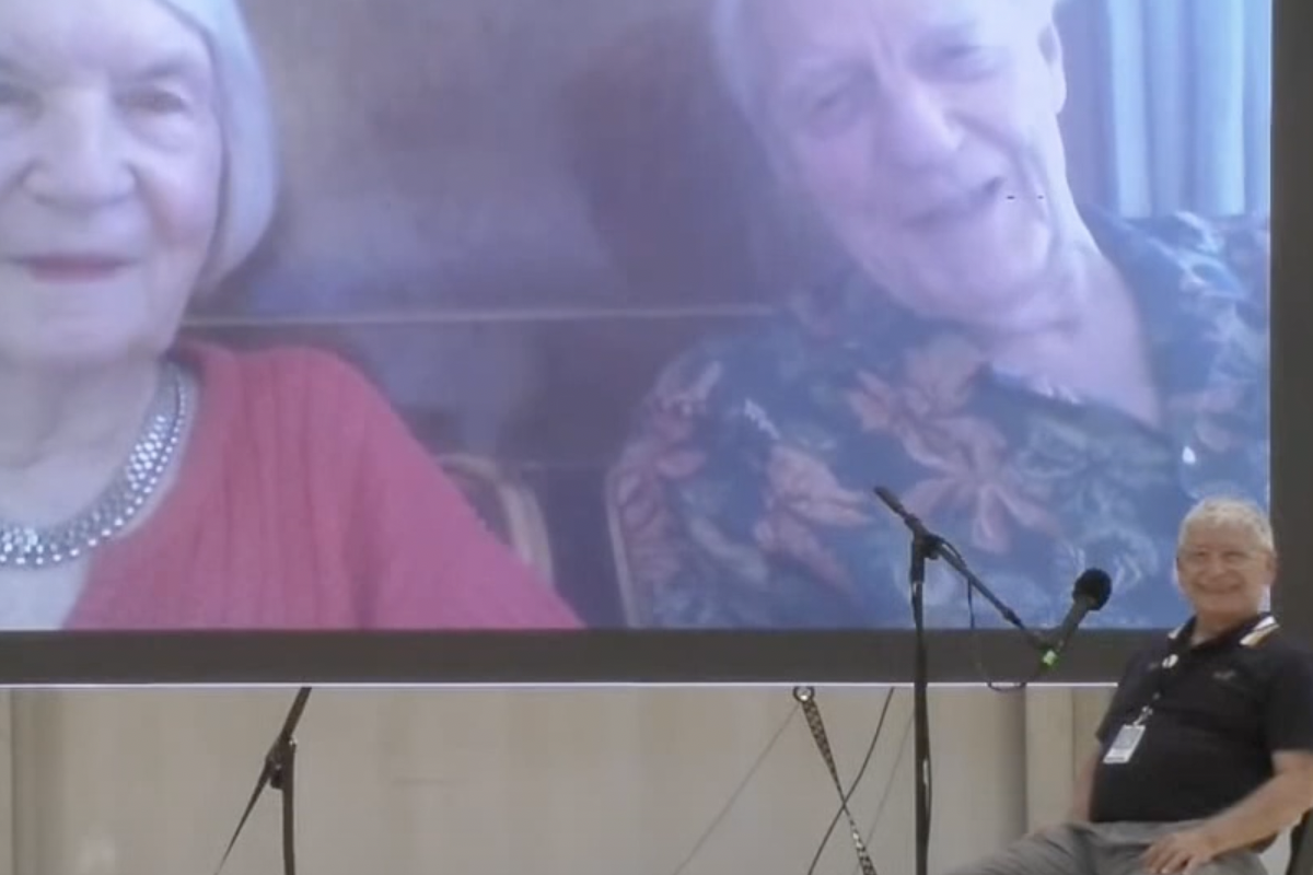 A smiling man sits in a chair facing the camera while two individuals, both slightly out of frame, appear on a large screen behind him.