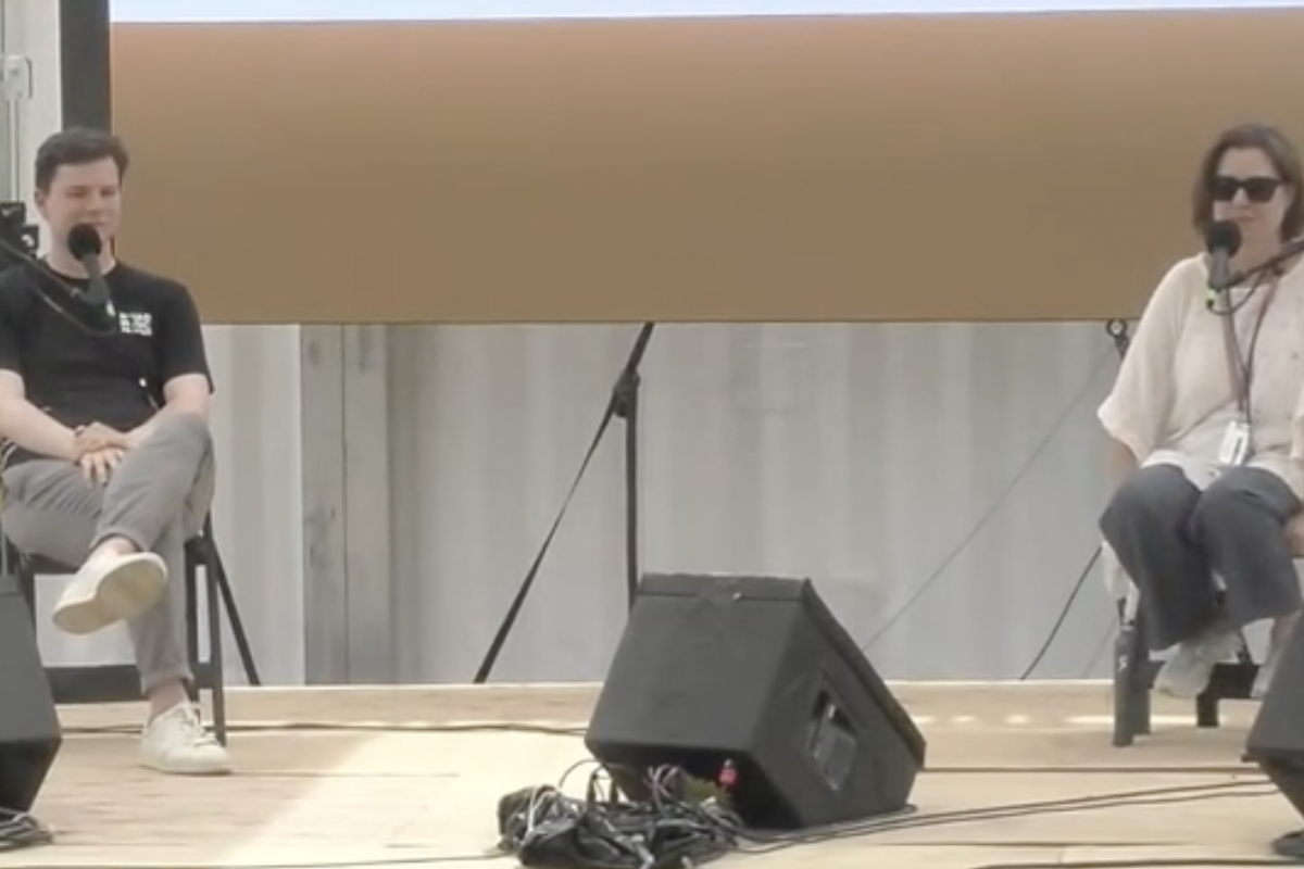 Two individuals sit on a stage, talking into microphones on stands.