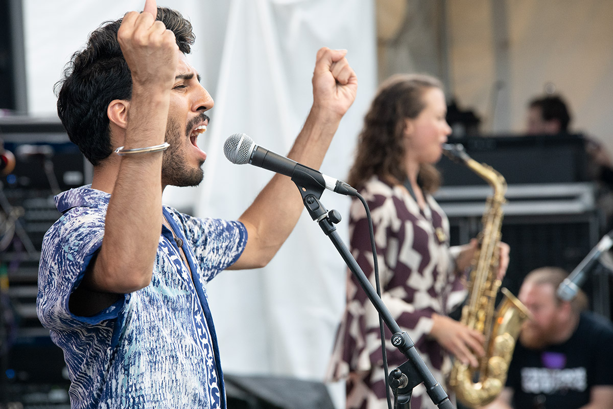 Two people performing on stage: a man singing, holding arms up in the air, and a woman playing saxophone.