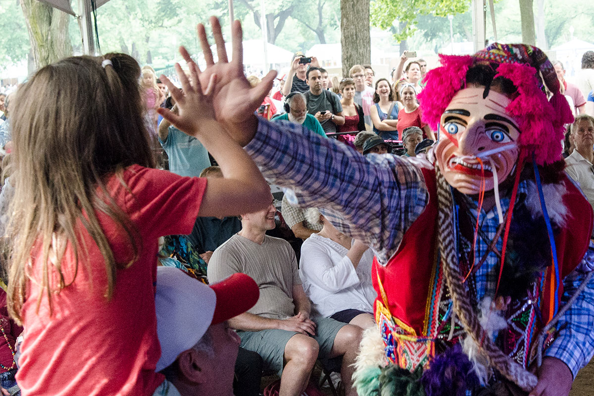 A Peruvian masked dancer high-fives a young child sitting on their father’s shoulders. A crowd of people watches in the background.