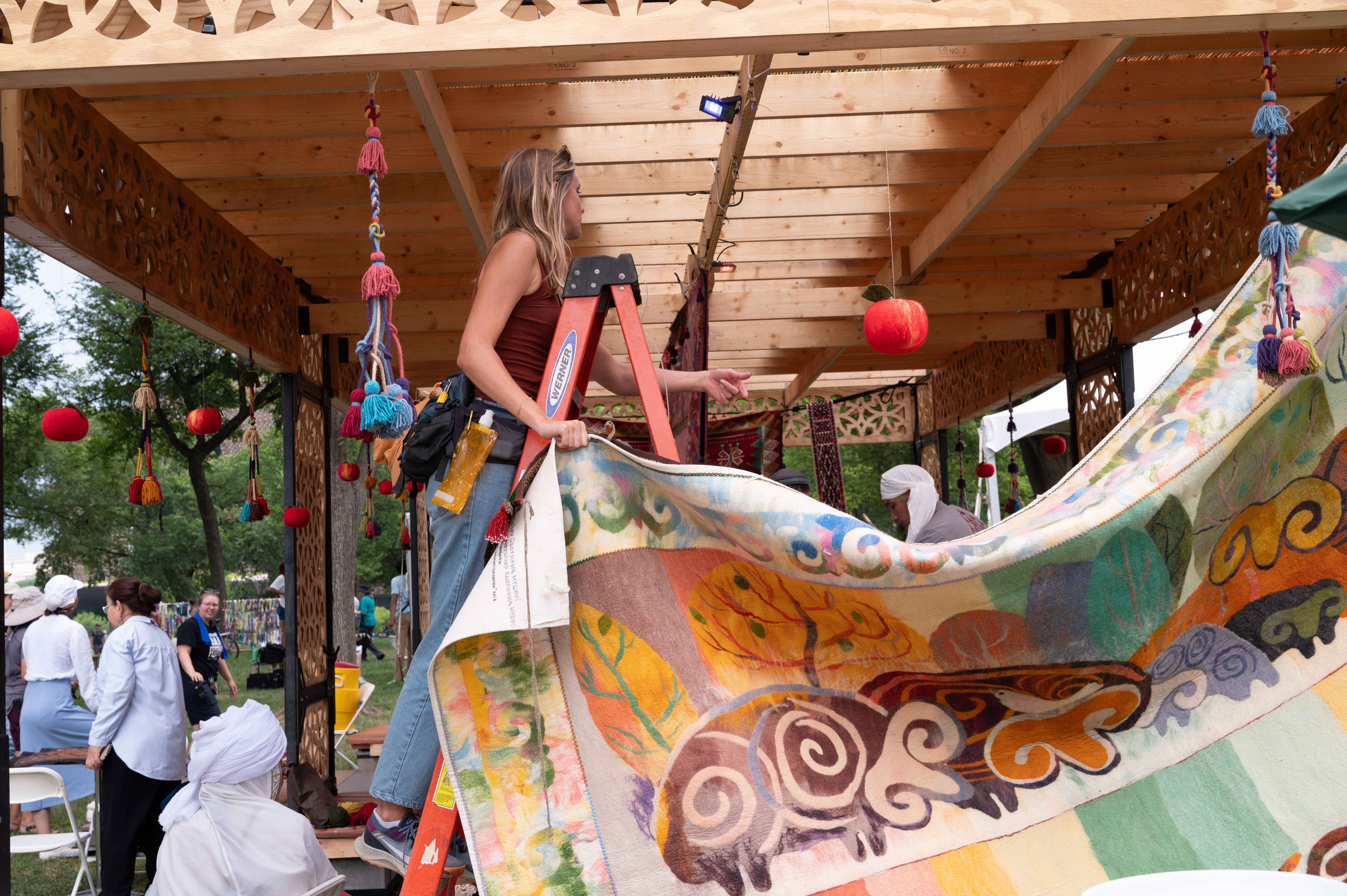 A woman on a ladder stands underneath a wooden building, holding the end of a tapestry depicting animals and trees in warm pastel colors, pointing off in the distance.