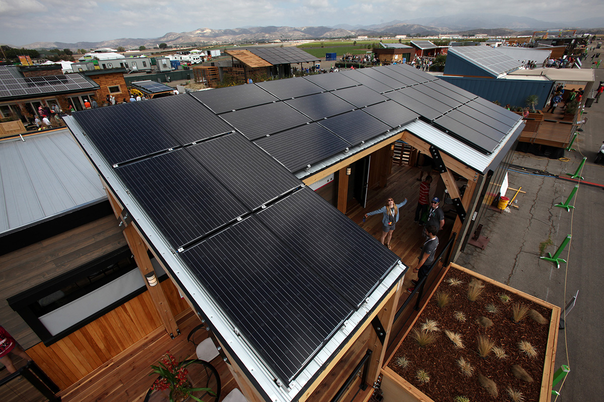 Aerial shot of a model home, with other models and mountains in the background. The roof of the house in focus is covered almost entirely in black solar panels. In the center, an open-air patio, is a woman looking up with outstretched arms.