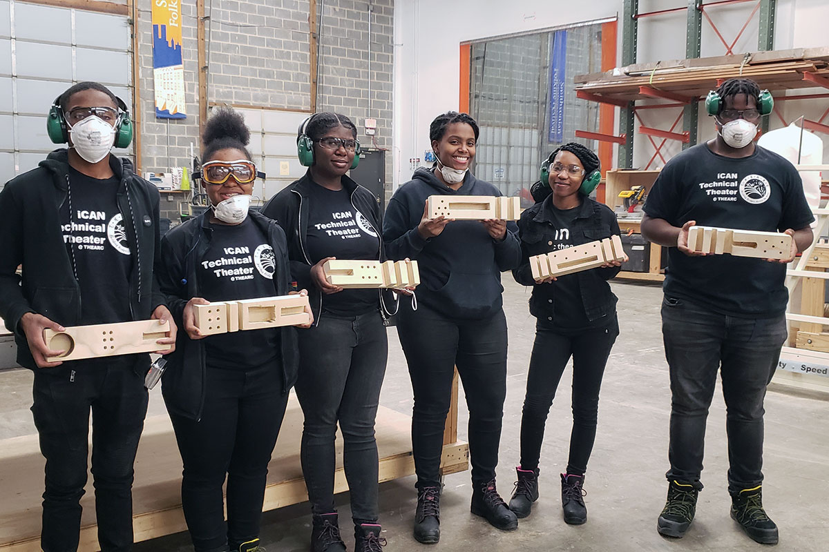 A group of teens in matching black outfits hold up wooden carpentry projects.