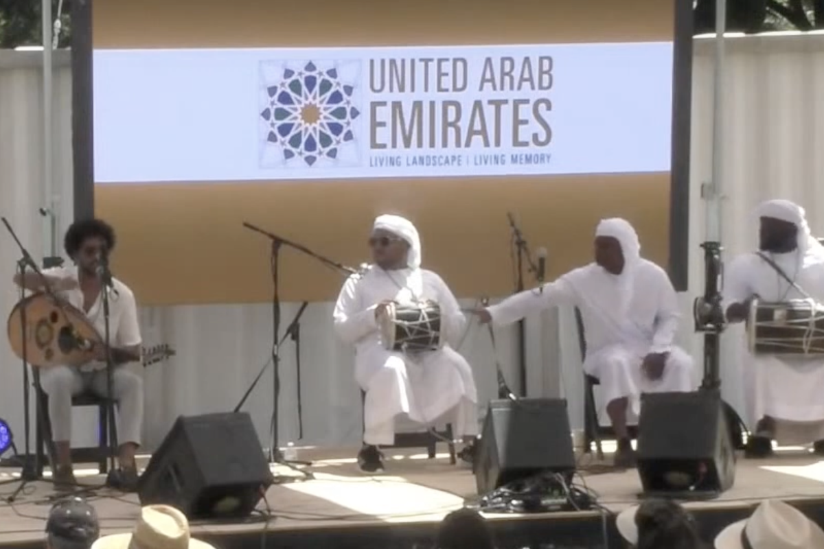 Four men seated on a stage holding various instruments, performing into microphone stands. Behind them, a screen reads "United Arab Emirates, Living Landscape, Living Memory."