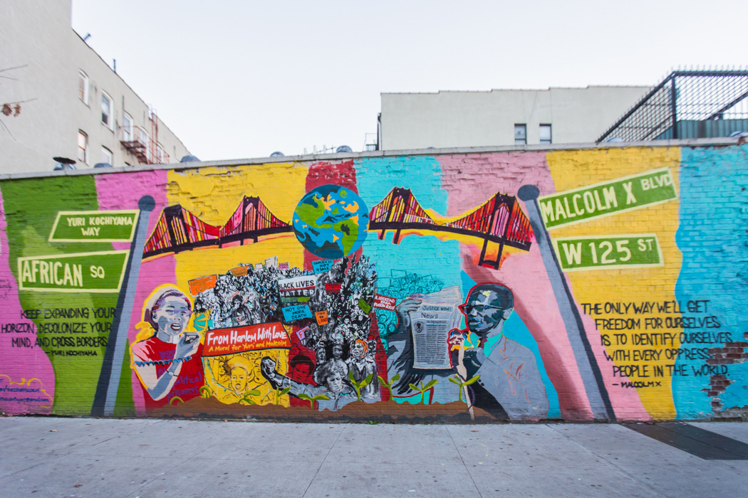 A brightly colored mural on a brick wall depicts movement leaders Yuri Kochiyama and Malcolm X, and speaking to crowds of protesters. On the left, a quote from Yuri: 'Keep expanding your horizon, decolonize your mind, and cross borders.' On the right, a quote from Malcolm: 'The only way we'll get to freedom for ourselves is to identify ourselves with every oppressed people in the world.' Between them, a bridge connects street signs for Yuri Kochiyama Way and Malcolm X Blvd., with a globe in the middle.
