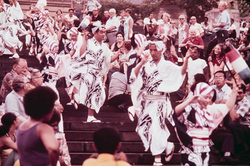 A group of people in white and black outfits dance down the stairs of stadium-style seating, with people seated on either side watching.