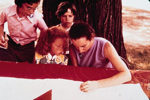A person hand-sews a red and white quilt outdoors, with three people watching closely.