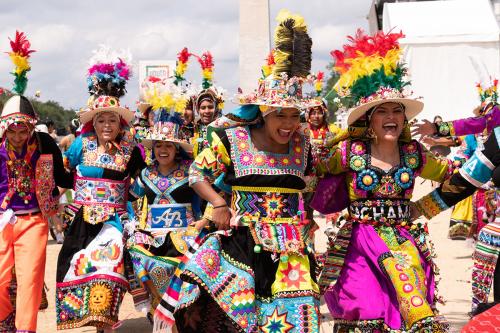 A group of smiling performers wearing colorful, ornate traditional costumes with large feather headdresses dancing in a festival parade.