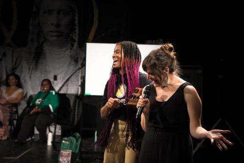Two women, one with pink locs and one with bangs and bun, laugh as they speak into a microphone to an audience.