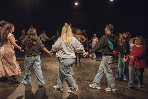 More than a dozen people form a circle in a dance room, holding hands and stepping forward. A woman at the center smiles, gesturing with one hand.