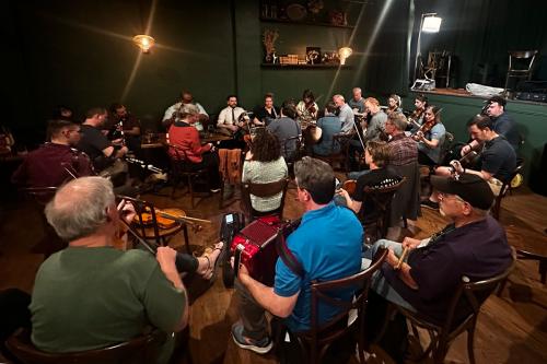 Two dozen people play various musical instruments, seated in loose concentric circles in a large room. There are fiddles, an accordion, guitars, and clarinets.